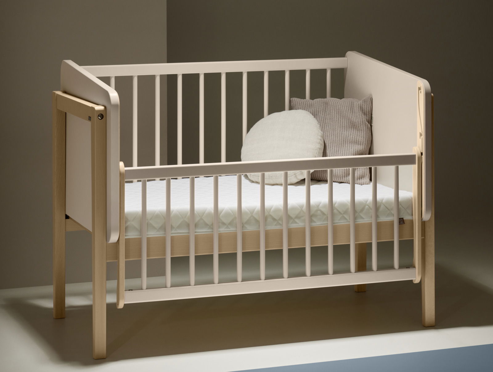 Anstellbett aus Holz mit weißem Gitter, seitliche Perspektive, zwei Kissen auf der Matratze.