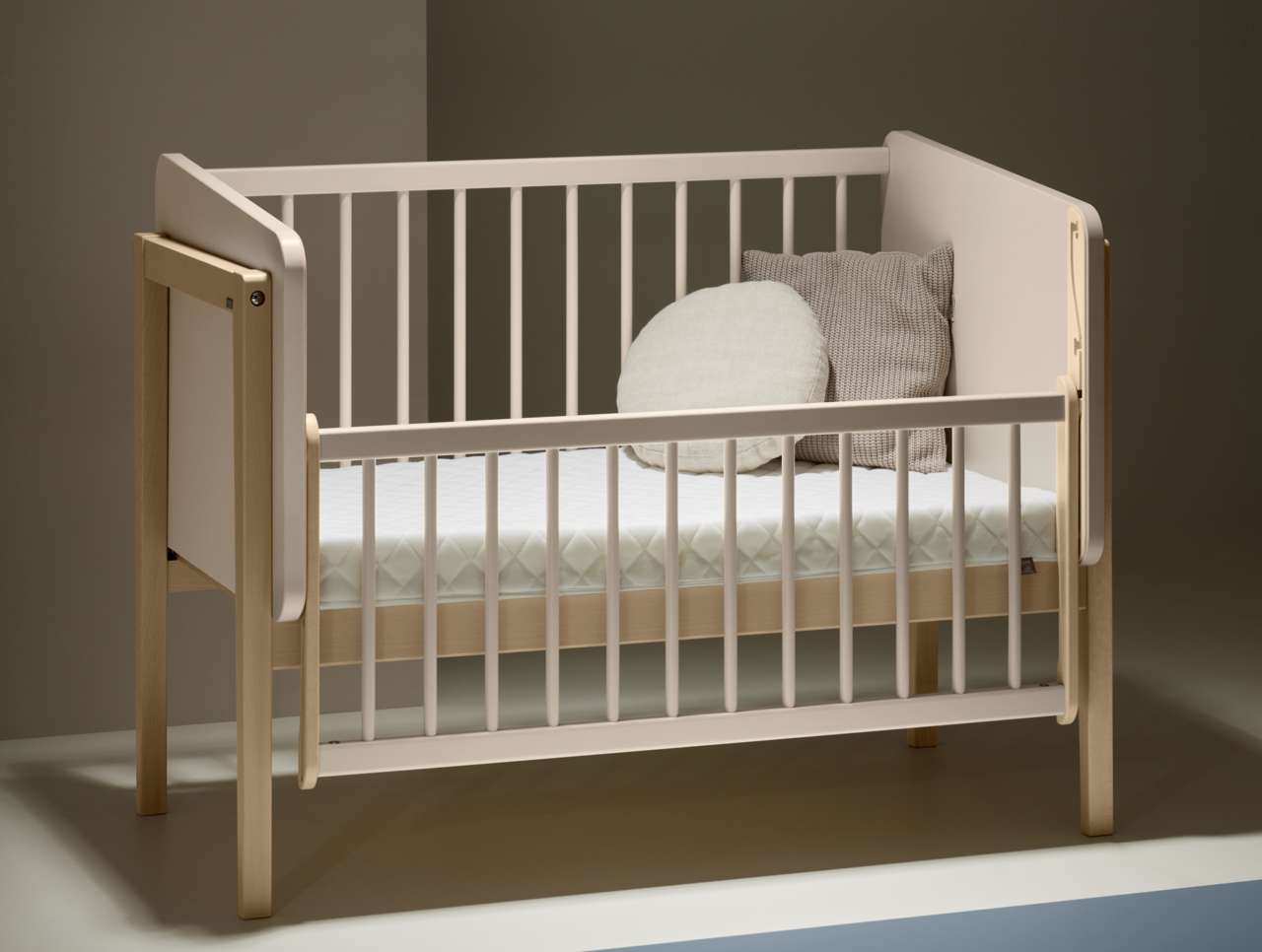 Anstellbett aus Holz mit weißem Gitter, seitliche Perspektive, zwei Kissen auf der Matratze.