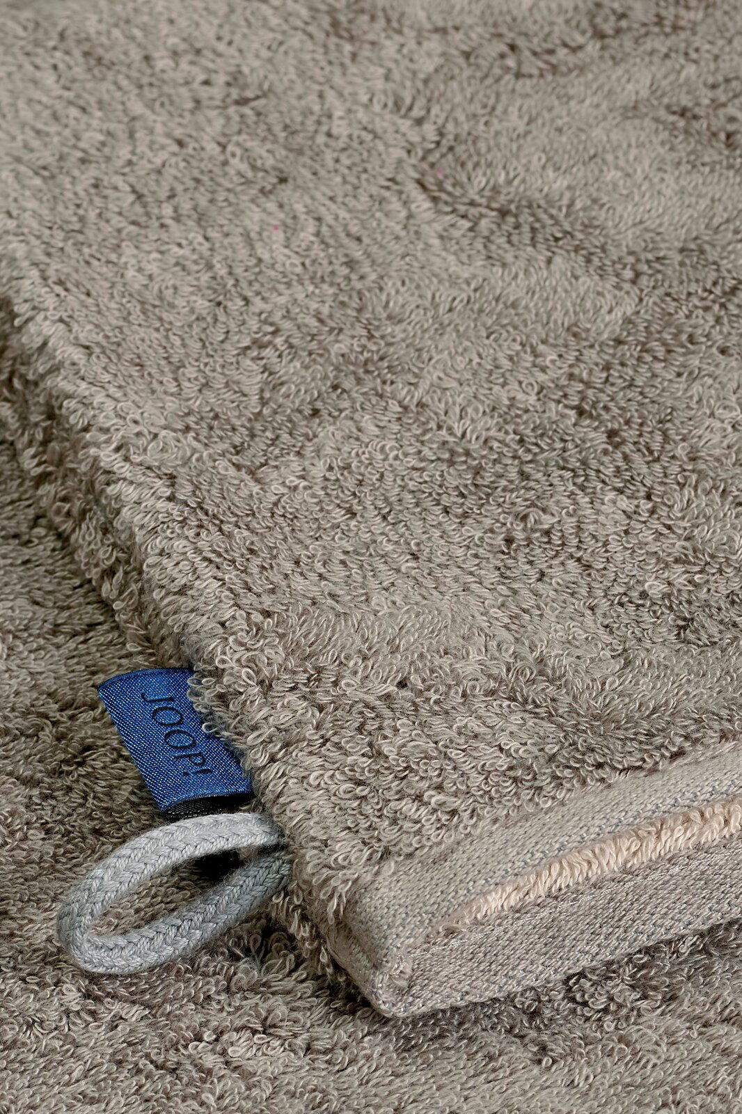 JOOP! Waschhandschuh DOUBLEFACE CLASSIC Nahaufnahme eines grauen Waschhandschuhs mit einer blauen Schlaufe und einem Etikett, liegend auf einer flauschigen Oberfläche.