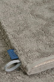 Nahaufnahme eines grauen Waschhandschuhs mit einer blauen Schlaufe und einem Etikett, liegend auf einer flauschigen Oberfläche.