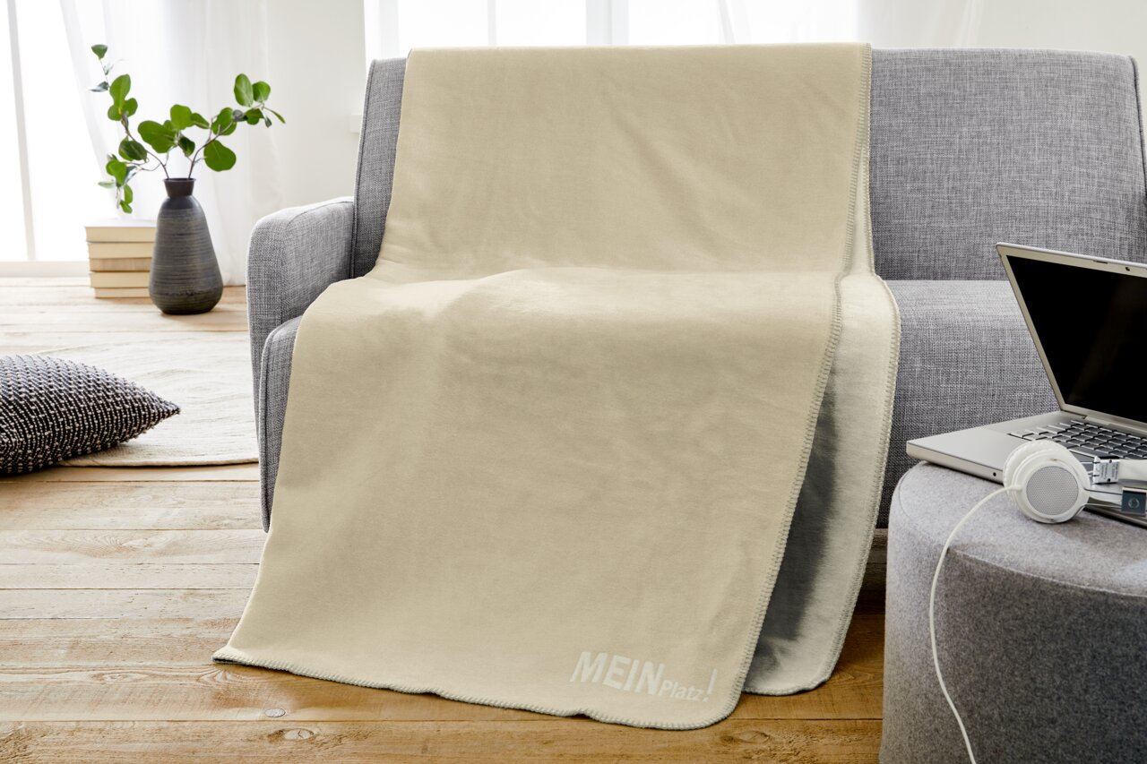Beige Jacquard-Decke mit der Aufschrift 'MEIN PLATZ!' auf einem grauen Sofa drapiert, seitliche Perspektive.
