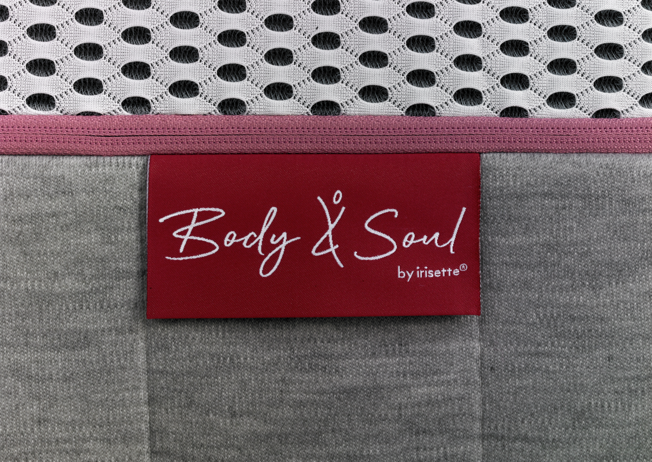 Nahaufnahme einer Matratze mit grauem Stoff und einem roten Etikett mit der Aufschrift 'Body & Soul by irisette'.