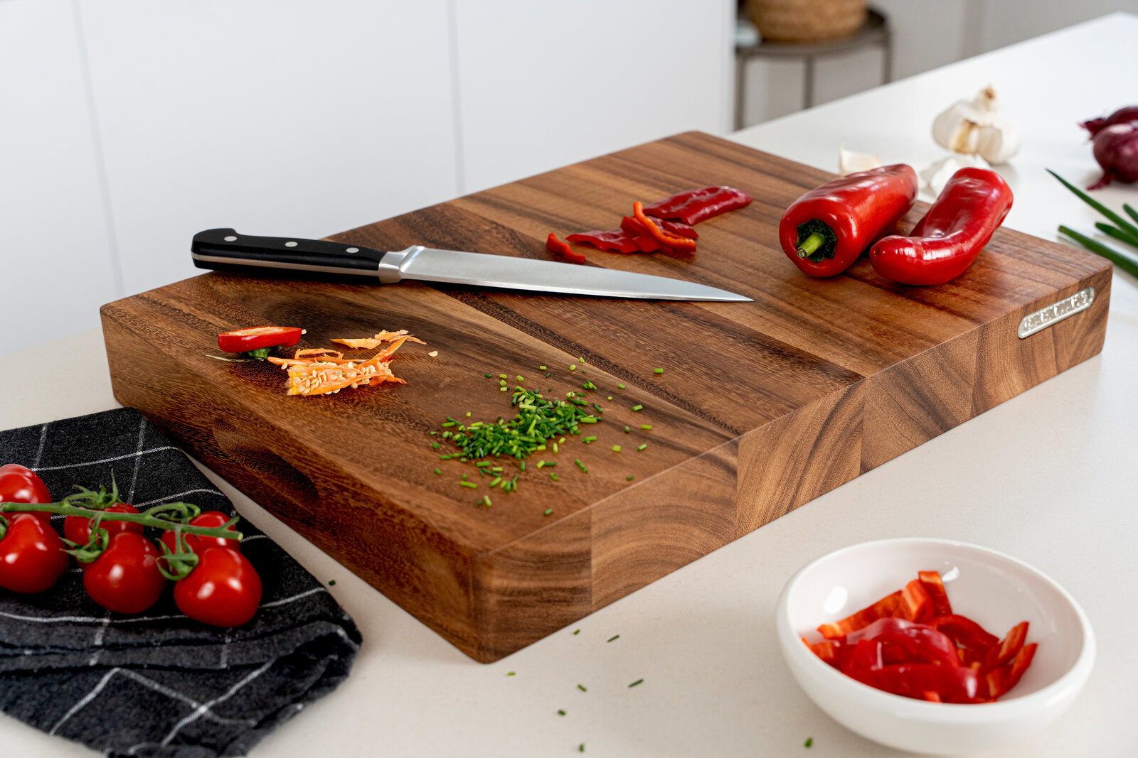 Massives Holz-Schneidebrett in der Küche mit Kochmesser, gehacktem Schnittlauch, Paprika und Tomaten, fotografiert aus schräger Seitenperspektive.