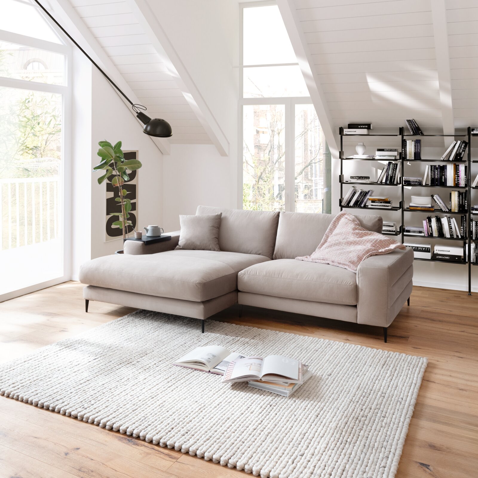 Beige Longchairkombination in einem modernen Wohnzimmer mit schrägen Decken, einem großen Fenster, einem Bücherregal und einem Teppich. Perspektive von vorne links.