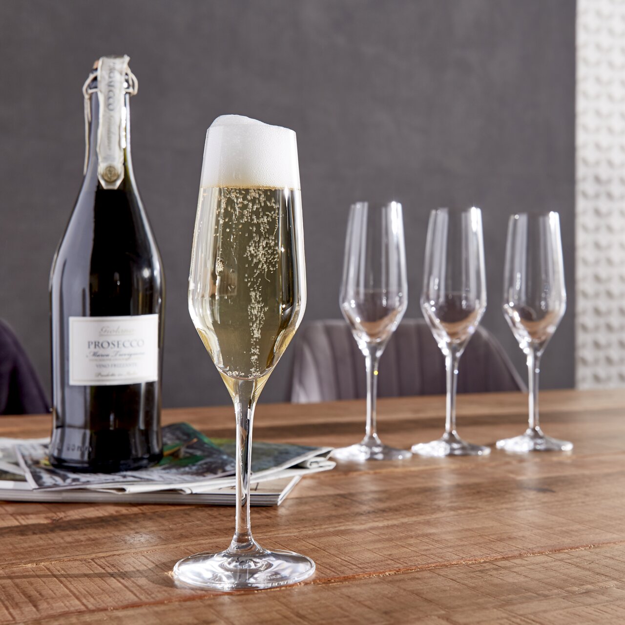 Spiegelau Champagnerkelch Style 4er-Set Ein Set aus vier eleganten Champagnerkelchen auf einem Holztisch, mit einer gefüllten Flöte im Vordergrund und einer Prosecco-Flasche daneben, seitliche Perspektive.