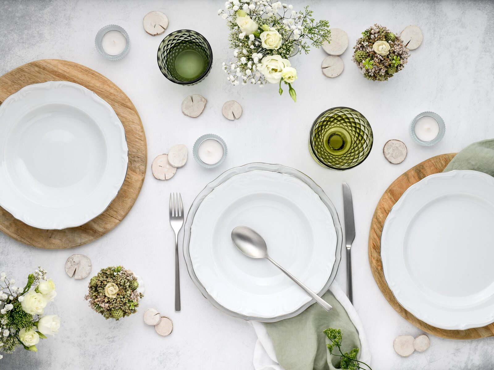 Draufsicht auf ein elegant gedecktes Tischarrangement mit weißen Porzellantellern auf runden Holzuntersetzern, grün getönten Gläsern, Besteck, Teelichtern und weißen Blumen auf hellem Hintergrund.