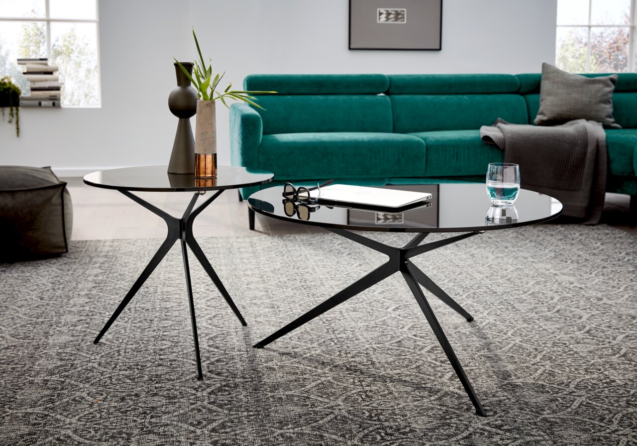 Zwei moderne Couchtische mit schwarzen Metallgestellen und runden Glasplatten, im Vordergrund von schräg oben fotografiert, vor einem grünen Sofa auf einem gemusterten Teppich.