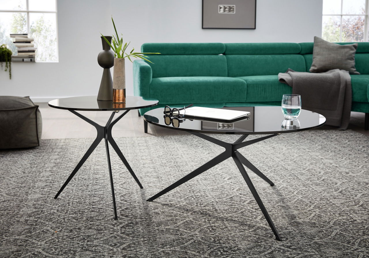 Zwei moderne Couchtische mit schwarzen Metallgestellen und runden Glasplatten, im Vordergrund von schräg oben fotografiert, vor einem grünen Sofa auf einem gemusterten Teppich.