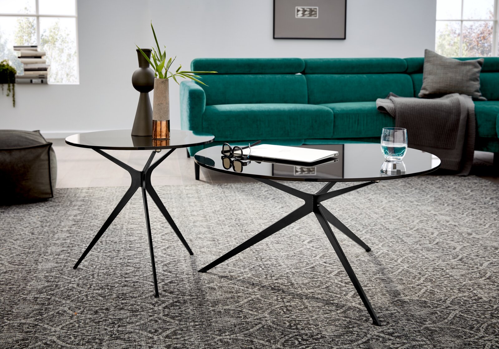 Twee moderne salontafels met een zwart metalen onderstel en ronde glazen bladen, op de voorgrond schuin van bovenaf gefotografeerd, voor een groene bank op een tapijt met een patroon.