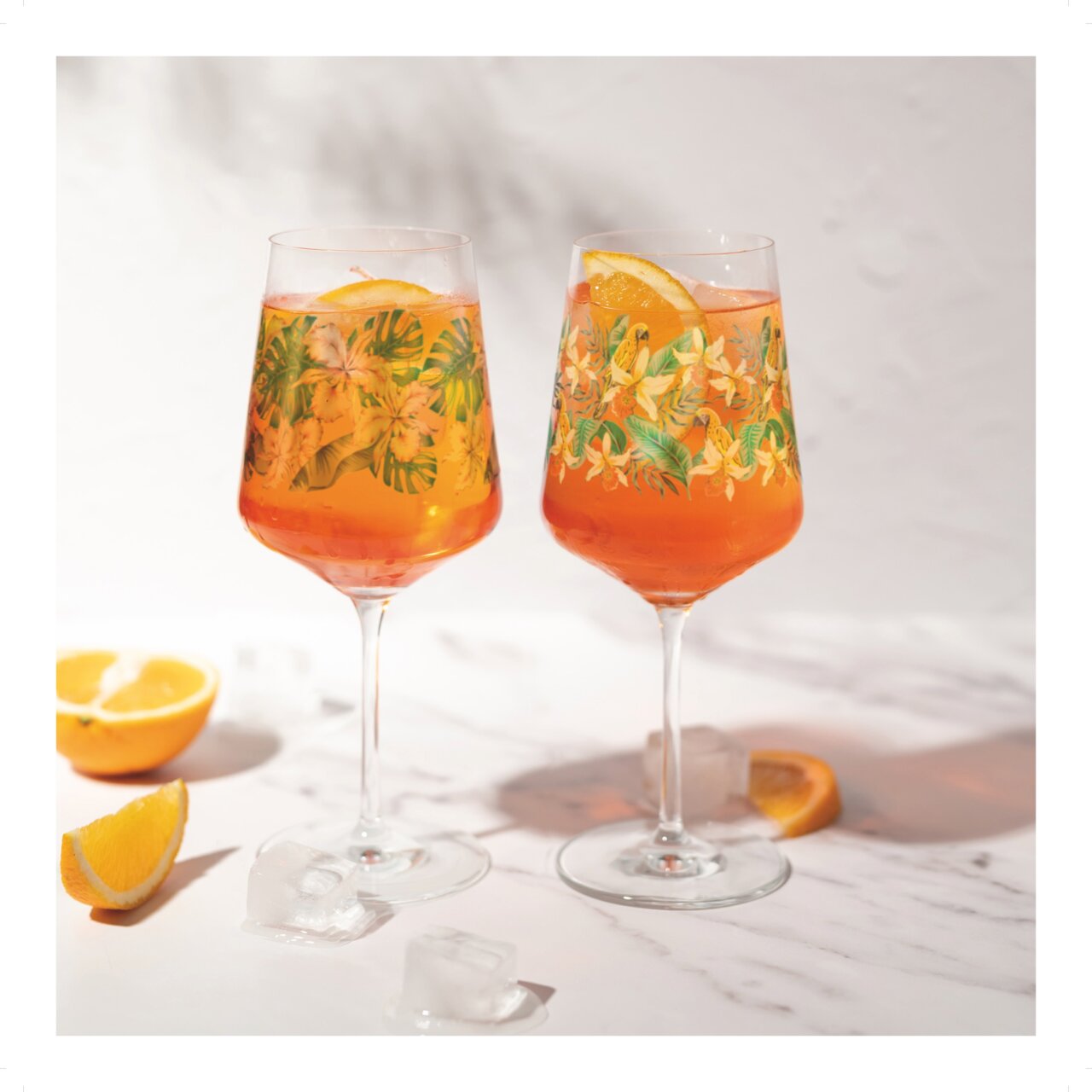 Zwei elegante Weingläser mit tropischem Blumenmuster, gefüllt mit einem orangefarbenen Getränk und Orangenscheiben, auf einer Marmoroberfläche mit Eiswürfeln und Orangenscheiben im Vordergrund, seitliche Perspektive.
