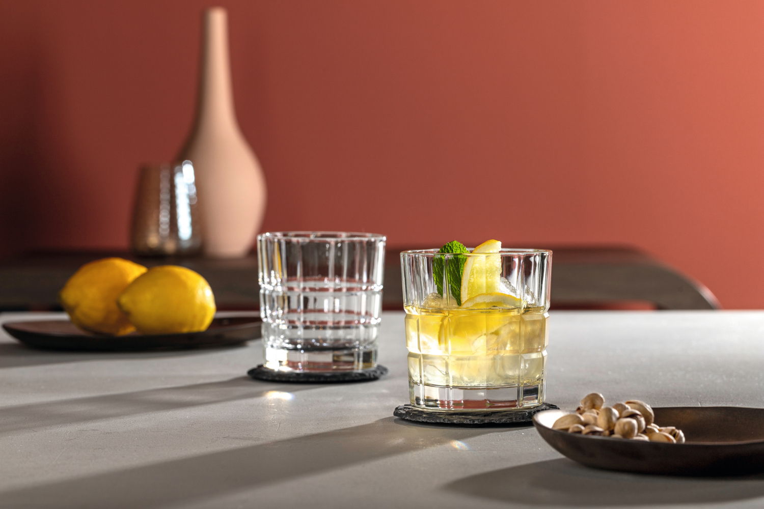 Zwei Whiskygläser SPIRITII 360 ml auf einem Tisch, eines gefüllt mit einem Getränk, Eiswürfeln und Zitronenscheiben, im Hintergrund eine Vase und Zitronen, seitliche Perspektive.