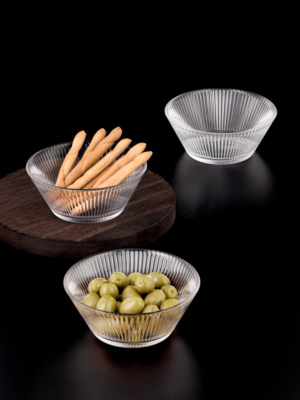 Drei Glasschalen mit geriffeltem Design, gefüllt mit Brotstangen und Oliven, auf schwarzem Hintergrund aus der Vogelperspektive.