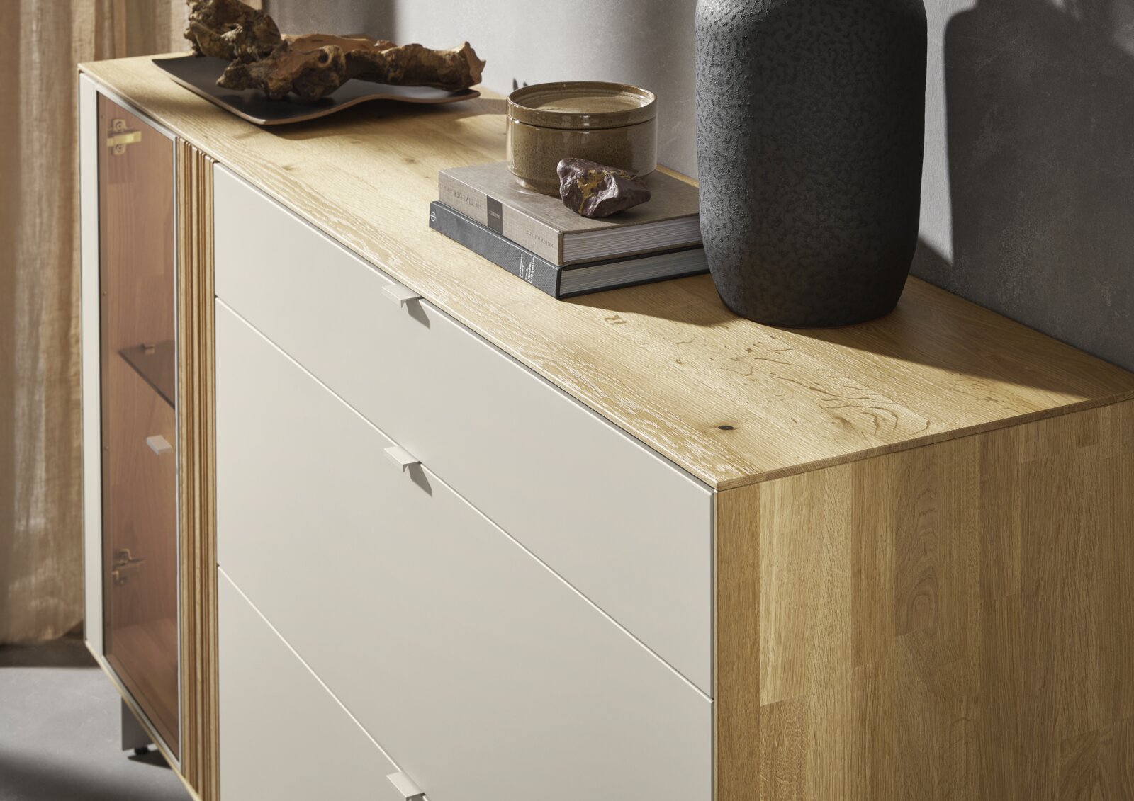 Moderne Kommode aus hellem Holz mit weißer Schubladenfront und seitlicher Glastür, dekoriert mit Büchern und Vase, in schräger Perspektive von vorne rechts aufgenommen.