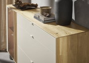 Moderne Kommode aus hellem Holz mit weißer Schubladenfront und seitlicher Glastür, dekoriert mit Büchern und Vase, in schräger Perspektive von vorne rechts aufgenommen.