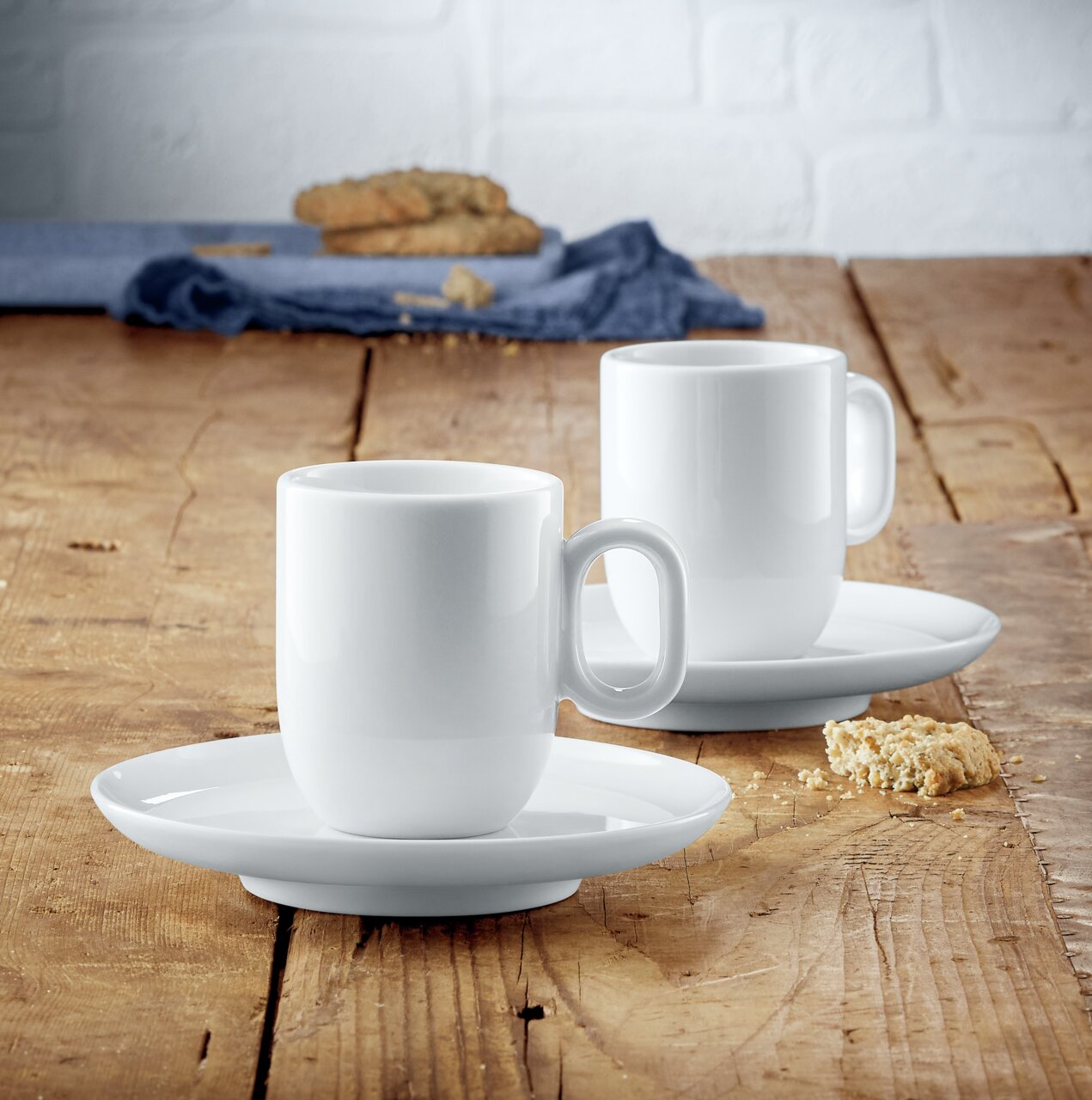 Zwei weiße Espressotassen mit Untertassen aus dem WMF Barista Espresso Tassen-Set auf einem Holztisch, seitliche Perspektive.