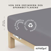 Graues Spannbettlaken aus Bio-Baumwolle und Elasthan auf einem Bett, mit Recycling-Verpackung, seitliche Perspektive.