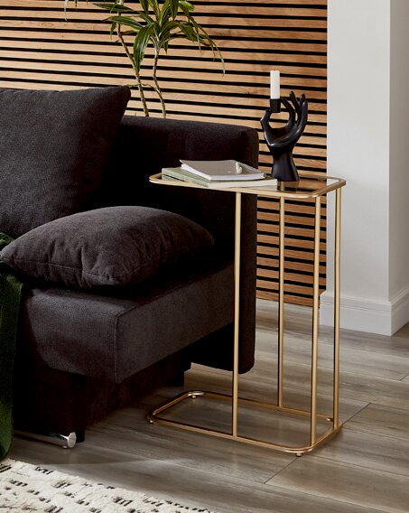 Seitliche Perspektive: Moderner Beistelltisch in Goldoptik mit rechteckigem Rahmen und Ablage neben dunkelgrauem Sofa, dekoriert mit Büchern und schwarzem Kerzenhalter vor Holzlamellenwand.