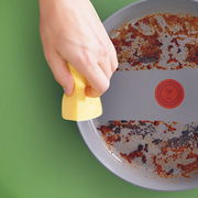 Hand reinigt eine Pfanne mit einem gelben Schwamm, Draufsicht auf die Pfanne mit eingebrannten Essensresten.