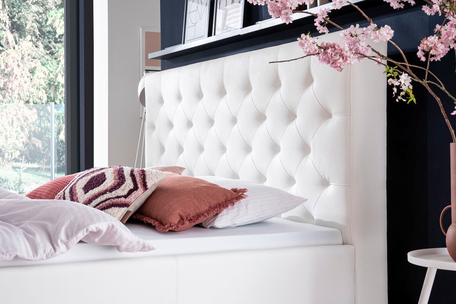 Seitliche Perspektive auf ein elegantes Polsterbett mit weißem, gestepptem Kopfteil, dekoriert mit Kissen und blühenden Zweigen.