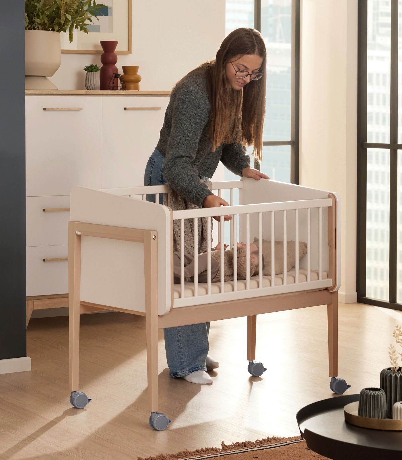 Frau steht neben einem Babybett mit Rollen in einem modernen Raum, seitliche Perspektive