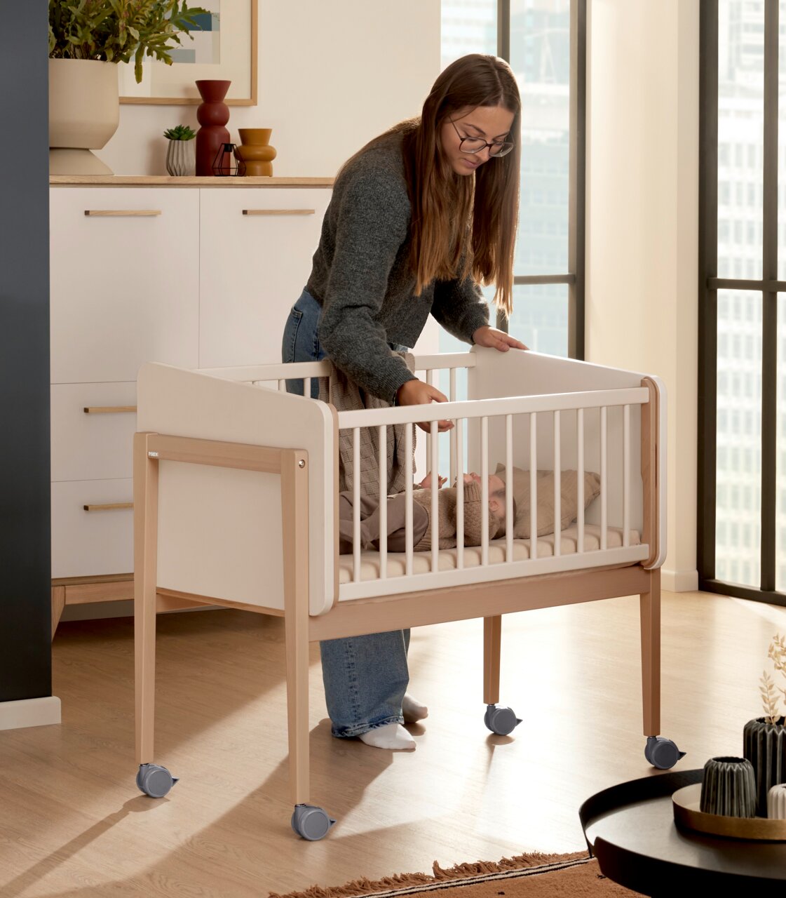 Een vrouw staat naast een wieg op wieltjes in een moderne kamer, gezien vanuit een zijdelingse hoek