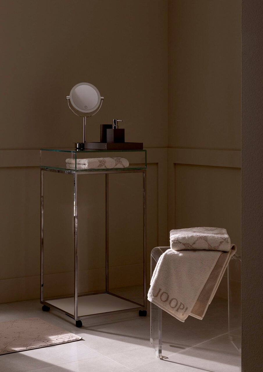 Een luxe handdoek in een stijlvolle badkamer, gedrapeerd over een doorzichtige stoel, met een elegant ontwerp en het opschrift 'JOOP!' in zijaanzicht.