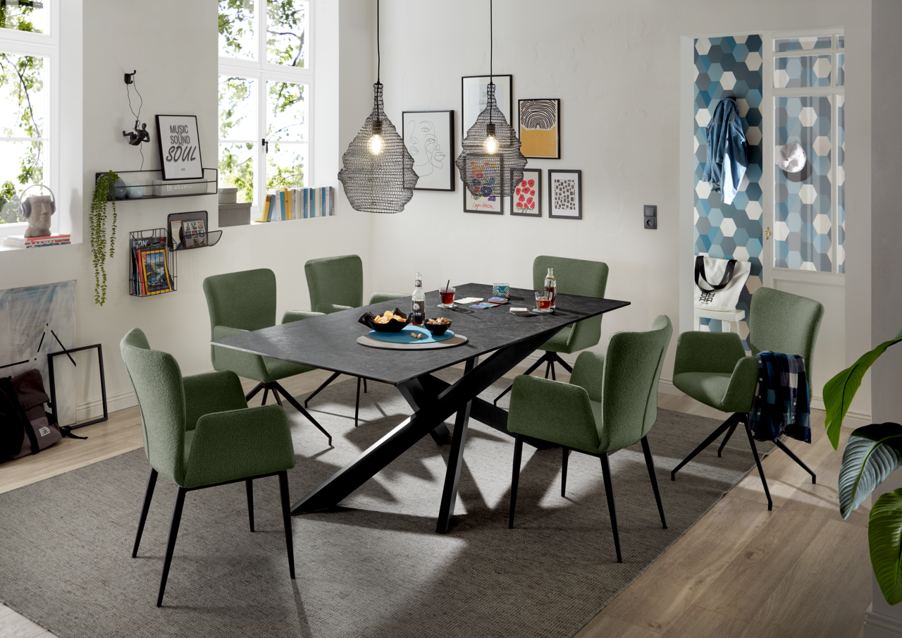 Moderner Esstisch mit schwarzer Tischplatte und X-förmigem Gestell, umgeben von grünen Stühlen in einem stilvoll eingerichteten Esszimmer, seitliche Perspektive.