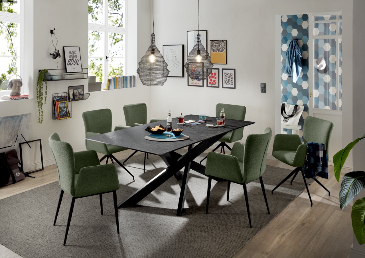 Moderner Esstisch mit schwarzer Tischplatte und X-förmigem Gestell, umgeben von grünen Stühlen in einem stilvoll eingerichteten Esszimmer, seitliche Perspektive.