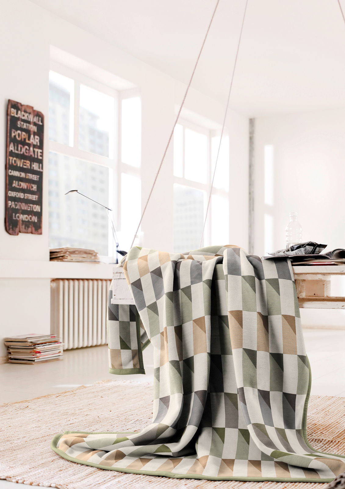 Gemusterte Wohndecke in Beige-, Grau- und Grüntönen, drapiert über einem Tisch in einem hellen Raum mit großen Fenstern.