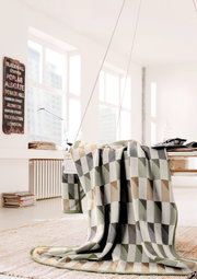 Gemusterte Wohndecke in Beige-, Grau- und Grüntönen, drapiert über einem Tisch in einem hellen Raum mit großen Fenstern.