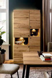 Frontale Ansicht einer modernen Wohnzimmervitrine aus hellem Massivholz mit zwei beleuchteten offenen Fächern und grifflosen Türen, im Wohnraum neben Fenster und Vorhang