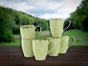 Fünf gesprenkelte Keramik-Kaffeebecher in hellgrün auf einem Holztisch vor unscharfem Waldhintergrund, leicht von vorne in Augenhöhe fotografiert