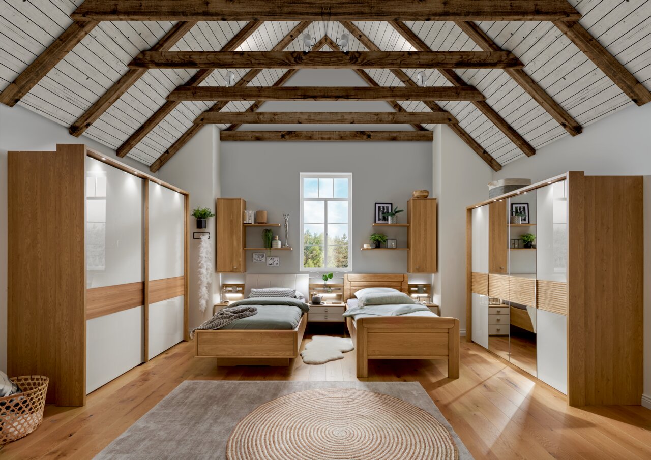 Schlafzimmer mit zwei Komfortbetten, umgeben von Schränken und Regalen in einem Raum mit Holzbalkendecke, aus zentraler Perspektive aufgenommen.