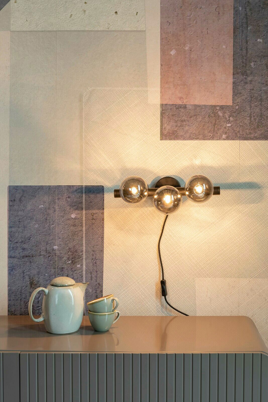 Moderne Wandleuchte mit drei kugelförmigen Glühbirnen, seitliche Perspektive, montiert über einem Tisch mit Teekanne und Tassen.