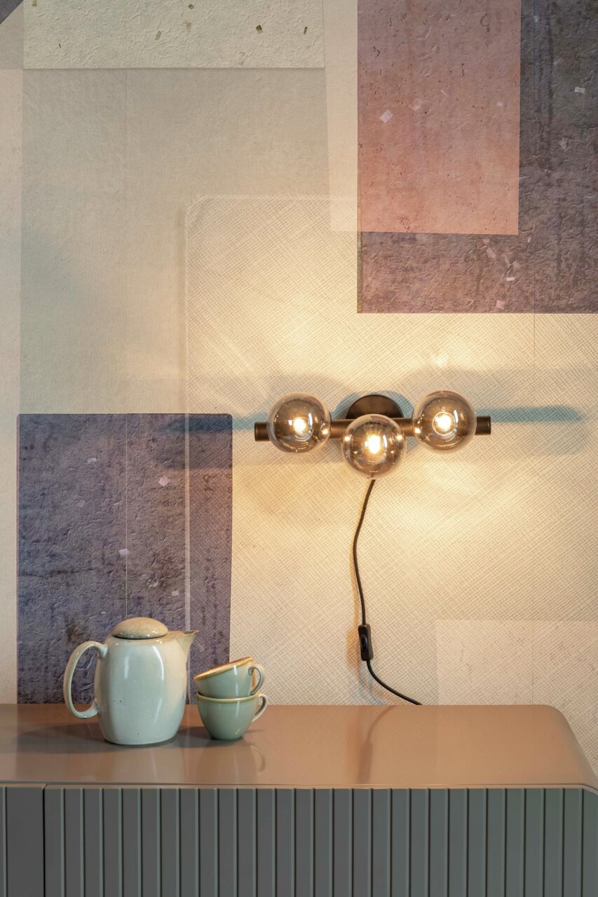 Moderne wandlamp met drie bolvormige lampen, zijaanzicht, bevestigd boven een tafel met een theepot en kopjes.