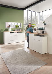 Moderner Wohnraum mit weißem Sideboard und Wandregal, dekoriert mit Pflanzen und Büchern, im Vordergrund eine Frau in entspannter Pose. Perspektive: seitlich von vorne.