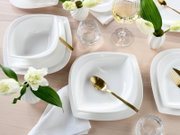 Draufsicht auf ein elegant gedecktes Tischarrangement mit weißen, quadratisch abgerundeten Porzellan-Tellern und tiefen Schalen, goldfarbenem Besteck, Wasser- und Weinglas sowie kleinen weißen Vasen mit Blumen auf hellem Holztisch.