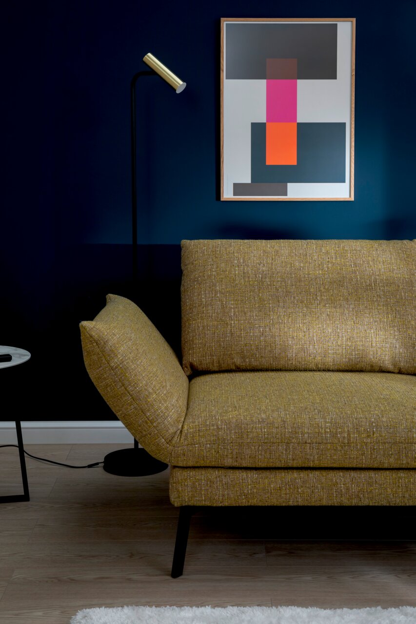 Gele longchair-bank in een moderne woonkamer, zijaanzicht met staande lamp en een abstract kunstwerk aan de muur.