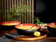 Schwarz-orangefarbene Schalen mit leuchtend gelber Innenseite, aus leicht erhöhter Frontperspektive auf einem runden Holztablett mit Limette und schwarzer Serviette arrangiert