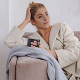 Frau in einem cremefarbenen Kapuzenbademantel, die auf einem Sofa sitzt und eine Tasse hält, seitliche Perspektive