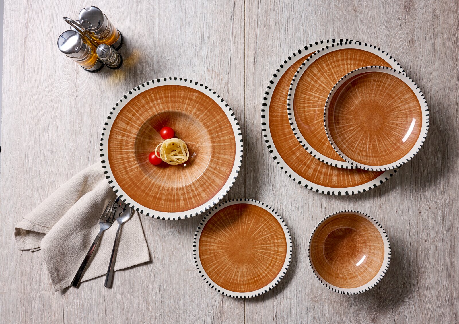 Bovenaanzicht van een rustiek servies met oranjebruine borden en kommen, witte randen en zwarte stipdetails op een lichte houten tafel