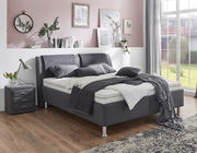 Modernes Polsterbett in einem stilvollen Schlafzimmer, seitliche Perspektive mit grauen Kissen und Decken, neben einem Nachttisch mit Lampe.