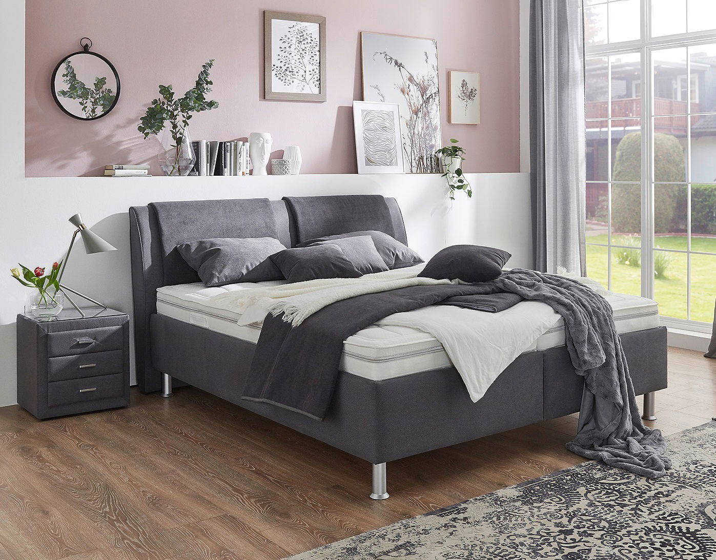 Modernes Polsterbett in einem stilvollen Schlafzimmer, seitliche Perspektive mit grauen Kissen und Decken, neben einem Nachttisch mit Lampe.