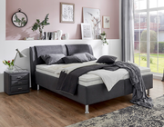 Modernes Polsterbett in einem stilvollen Schlafzimmer, seitliche Perspektive mit grauen Kissen und Decken, neben einem Nachttisch mit Lampe.