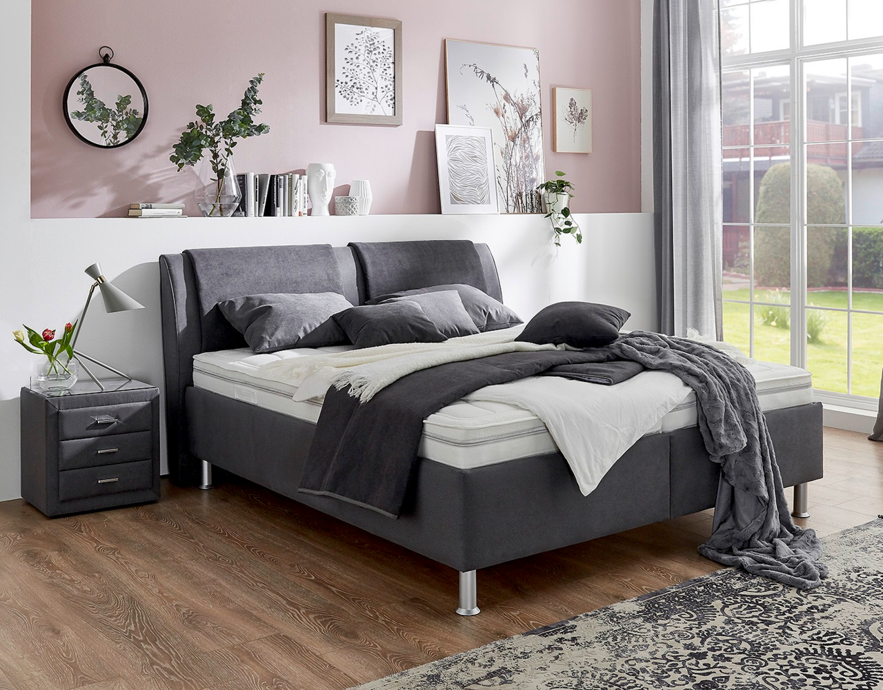 Modernes Polsterbett in einem stilvollen Schlafzimmer, seitliche Perspektive mit grauen Kissen und Decken, neben einem Nachttisch mit Lampe.