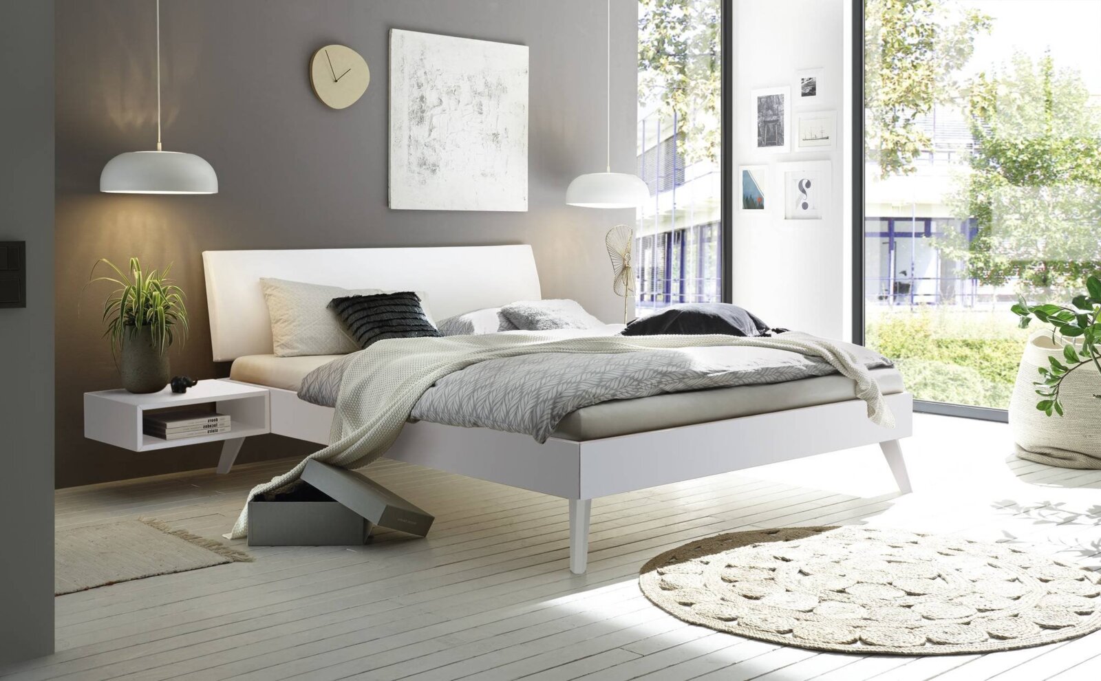 Modernes weißes Bettgestell in einem stilvollen Schlafzimmer mit grauer Wand, hängenden Lampen und großen Fenstern, seitliche Perspektive
