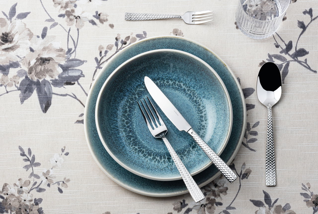 Vogelperspektive auf ein stilvolles Besteckset mit Messer, Gabel und Löffel auf blauer Keramik, umgeben von floraler Tischdecke.