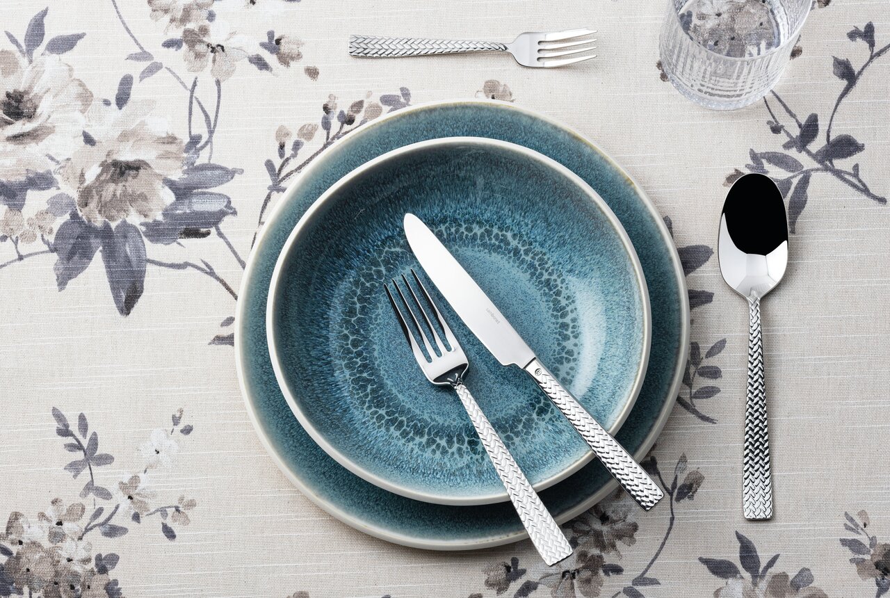 Vogelperspektive auf ein stilvolles Besteckset mit Messer, Gabel und Löffel auf blauer Keramik, umgeben von floraler Tischdecke.