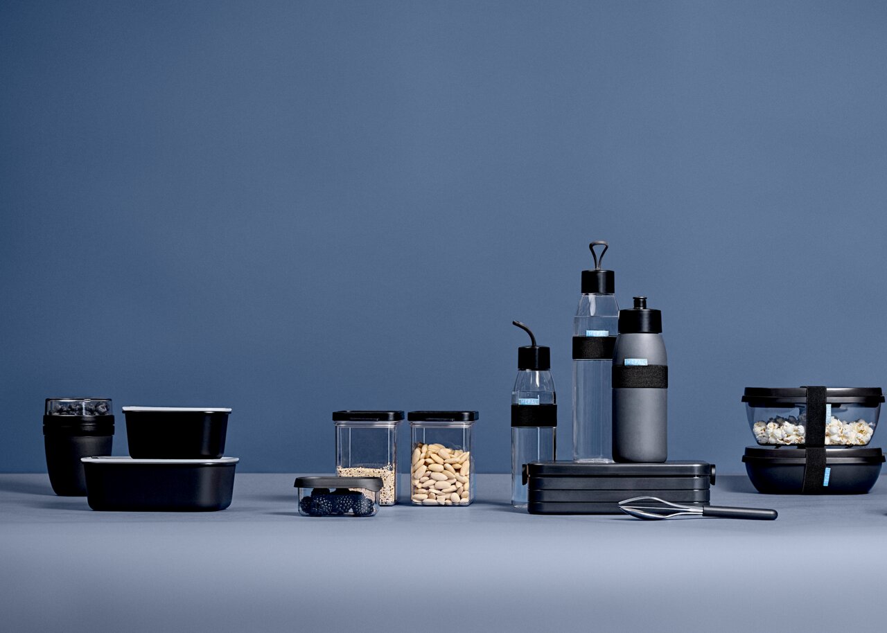 Verschillende zwarte bewaardozen en drinkflessen in een moderne, minimalistische opstelling tegen een blauwe achtergrond, gezien vanuit een zijdelingse hoek.