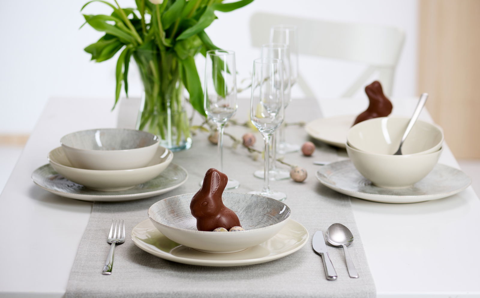 Elegantes 18-teiliges Geschirrset auf einem gedeckten Tisch mit Frühlingsdekoration, aus der Perspektive von vorne betrachtet.